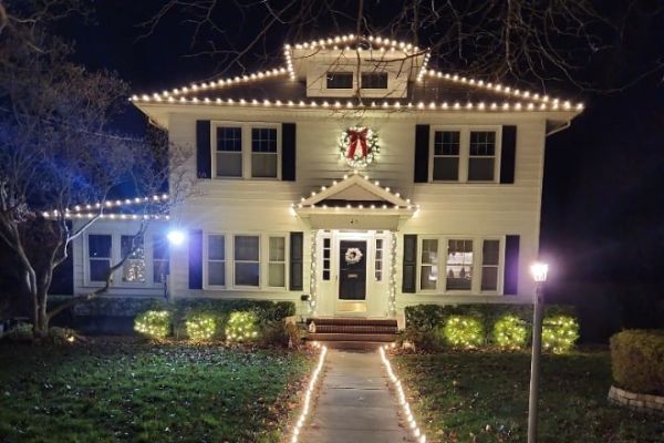 A charming house glows with Christmas lighting, featuring lights along the roofline, walkway, and bushes. Wreaths adorn the door and second-story window, adding to the festive holiday scene.