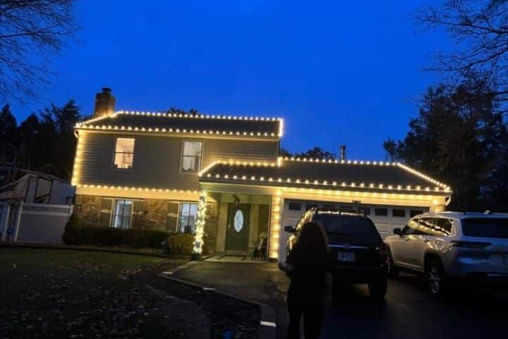 House adorned with warm white Christmas lighting along the roofline. Cars parked in the driveway, silhouetted figure in the foreground. Festive, inviting Christmas Lighting scene.