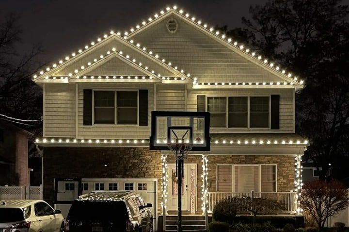 Two-story house adorned with bright white Christmas lighting outlining the roof, porch, and front door. A basketball hoop stands in front of the house, adding a touch of everyday life to the festive scene.