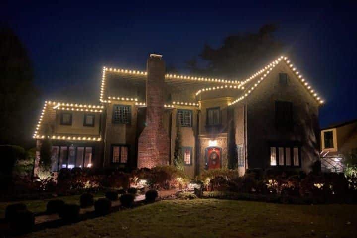 A large, stately house is decorated with warm white Christmas lights outlining the roofline and windows, creating a cozy and festive ambiance in the Gallery.